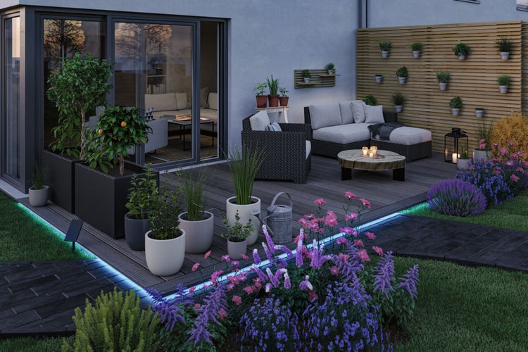 Moderne Holzterrasse bei Dämmerung mit Lichtstreifen, Gartenmöbeln aus Geflecht, verschiedenen Pflanzgefäßen und einem Sichtschutzzaun aus Holz.