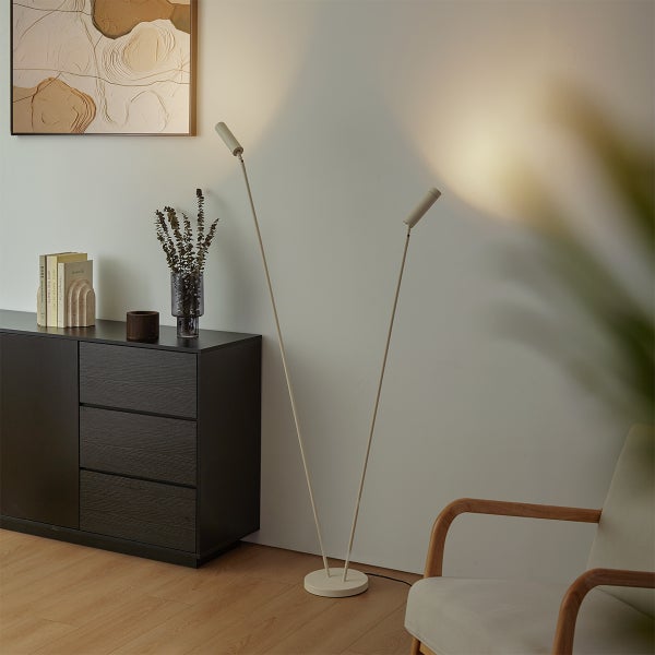 Moderne zweiflammige Stehleuchte in Beige in einem minimalistischen Wohnzimmer neben einer dunklen Kommode und einem Sessel.