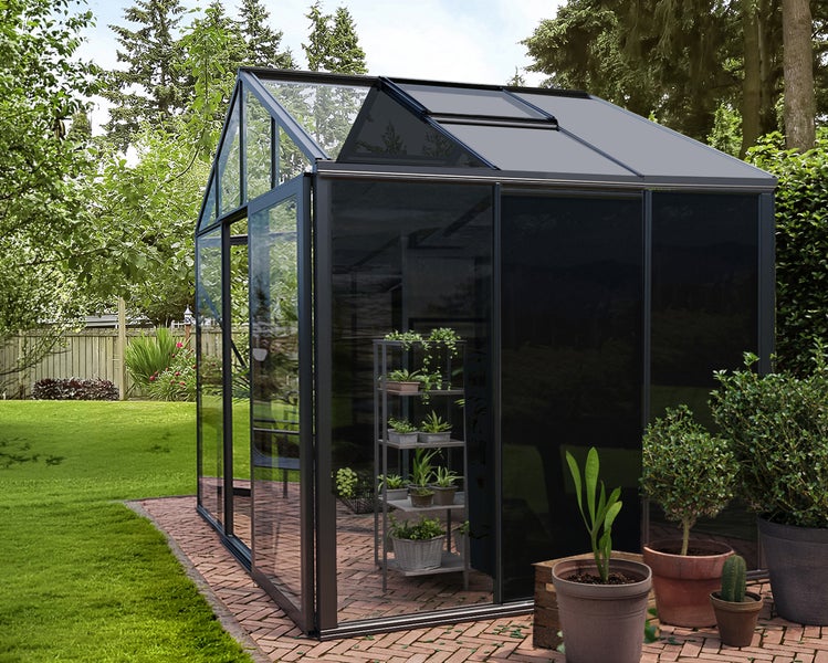 Gewächshaus mit dunklem Aluminiumrahmen und Glaspaneelen im Garten. Mit Schiebetür, Dachfenster und innenstehendem Regal auf einer Steinterrasse.