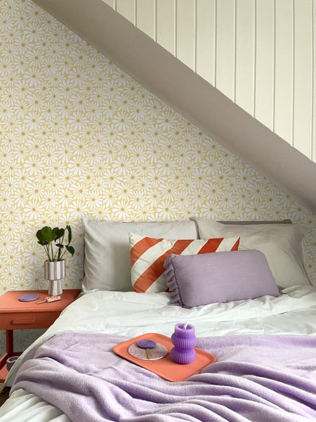 Helles Schlafzimmer im Dachgeschoss mit Gänseblümchen-Tapete, Bett mit Dekokissen und Wandverkleidung aus Holz.