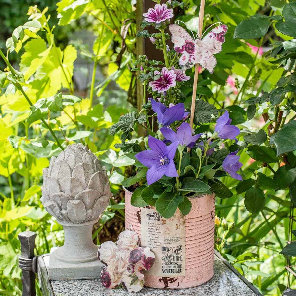 Gartendekoration mit lila Blumen im Blechtopf, einer Steinskulptur in Zapfenform und Schmetterlingsstecker inmitten grüner Pflanzen.