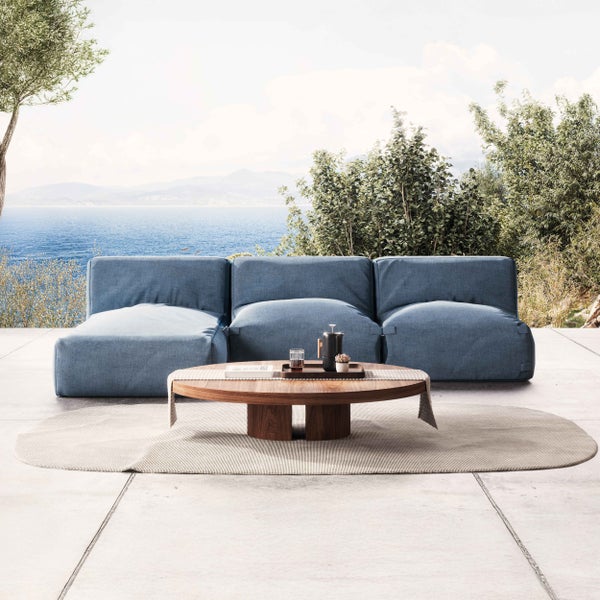 Moderner Outdoor-Loungebereich auf heller Terrasse mit modularem blauem Sofa, rundem Holztisch und ovalem Teppich mit Meerblick.