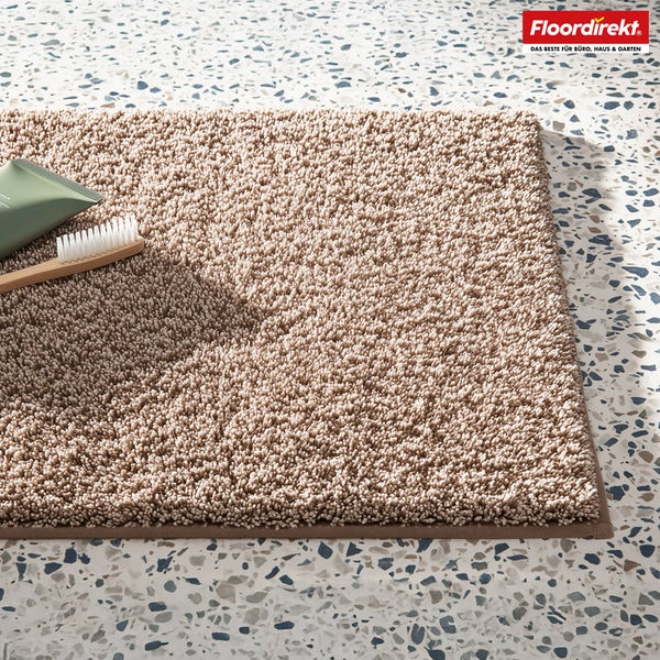 Beige Badematte mit Zahnbürste und Zahnpasta auf einem gefleckten Untergrund.