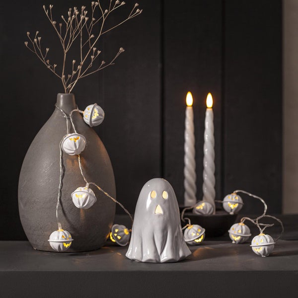 Dekorative Halloween-Szene mit einer Geisterfigur, einer Kürbis-Lichterkette und einer Vase mit Zweigen.