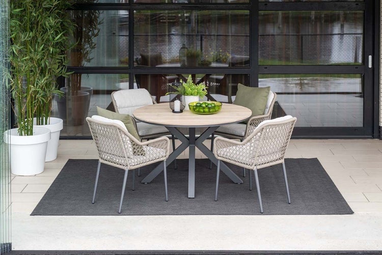Modernes Gartenmöbel-Set mit rundem Holztisch und vier Geflechtsesseln auf einem Außenteppich vor einer Glasfront.