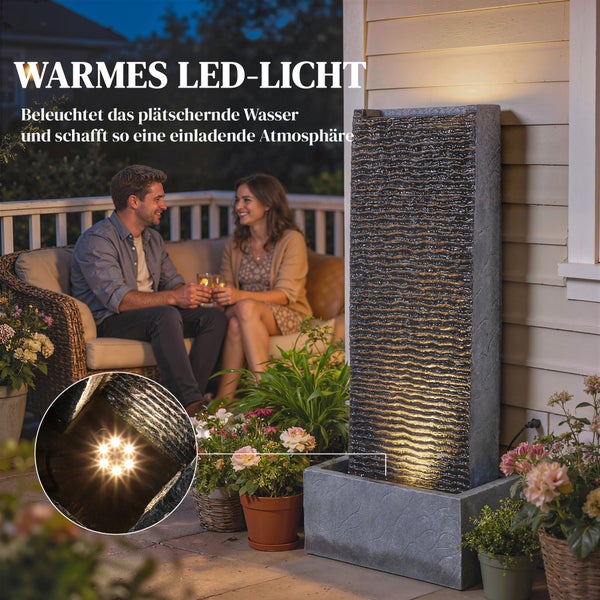Moderner Gartenbrunnen in Steinoptik mit warmem LED-Licht auf einer Terrasse bei Nacht, neben einer Sitzgruppe mit Personen und Topfpflanzen.