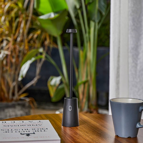 Moderne, schwarze LED Akku-Tischleuchte auf einem Holztisch neben einem Buch und einer Tasse vor grünem Hintergrund.