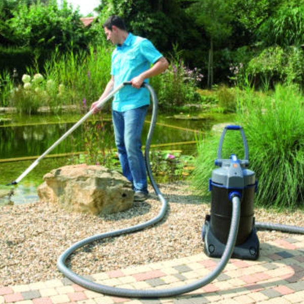 Ein Mann reinigt einen bepflanzten Gartenteich mit einem Teichsauger auf einem Kieselweg neben einer Steinmauer.