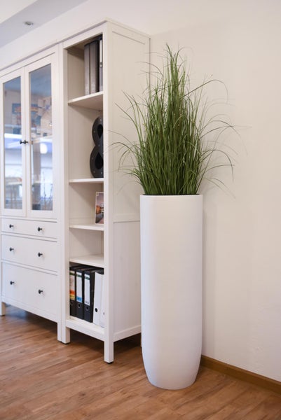 Hoher weißer runder Pflanzkübel mit künstlichem Gras neben einem weißen Regal in einem hellen Zimmer mit Holzboden.