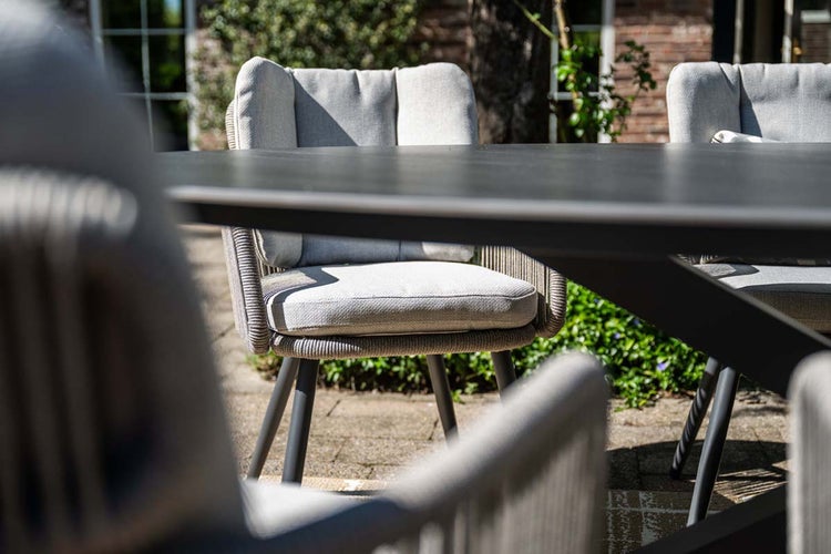 Moderner Gartenstuhl mit Kordelgeflecht und hellgrauen Sitzkissen an einem dunklen Gartentisch auf einer Terrasse.