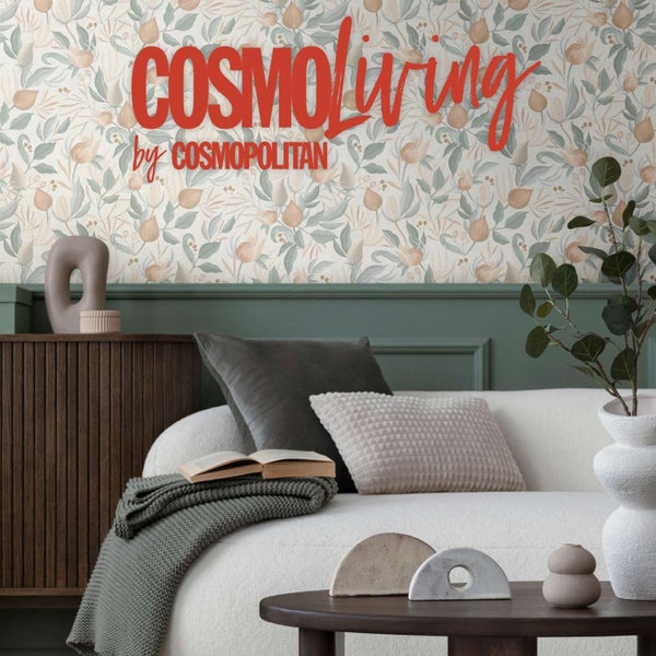 Wohnzimmer mit Vliestapete mit Blumenmuster, Cosmoliving by Cosmopolitan Logo, Sofa mit Dekokissen, Holzkommode und Zimmerpflanze in Vase.