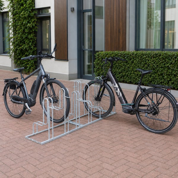 Fahrradständer aus Metall auf einem gepflasterten Vorplatz mit zwei abgestellten Elektrofahrrädern vor einem modernen Gebäude.