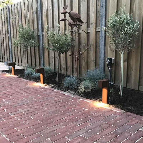 Gartenweg mit Pflastersteinen, Pollerleuchten in Rostoptik, Olivenbäumen und Holzzaun.