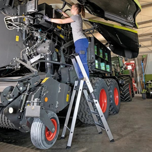 Eine Person steht auf einer stabilen Aluminium Stehleiter und führt Wartungsarbeiten an einer landwirtschaftlichen Maschine in einer Werkstatt durch.