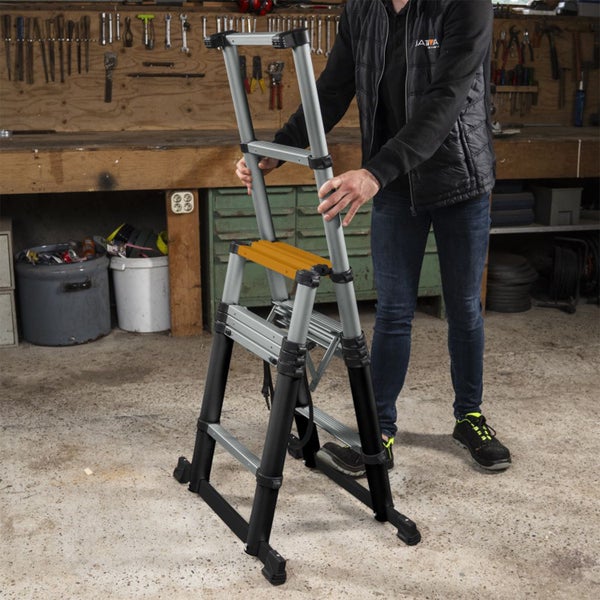 Eine Person hält eine silberne und schwarze Teleskop-Kombileiter aus Aluminium in einer Werkstatt mit Werkbank und Werkzeugwand.