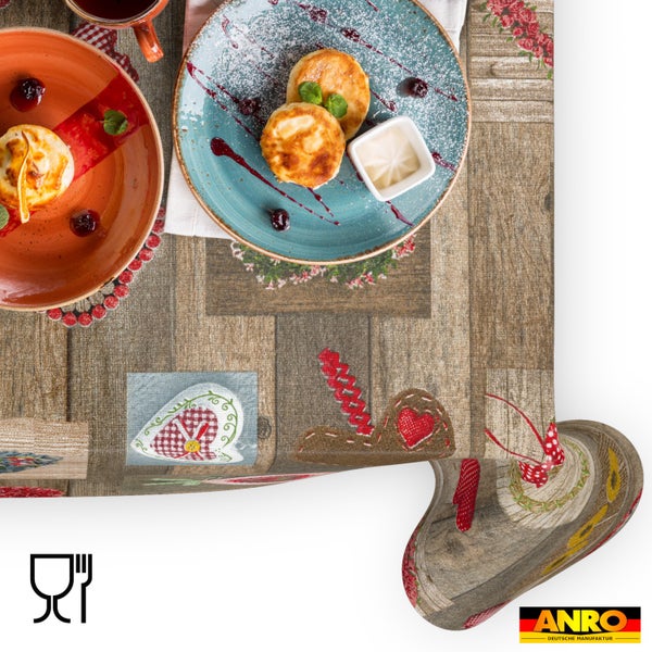 Abwaschbare Tischdecke in Holzoptik mit Herzmotiven, gedeckt mit Tellern. Lebensmittelecht. ANRO Deutsche Manufaktur Logo.