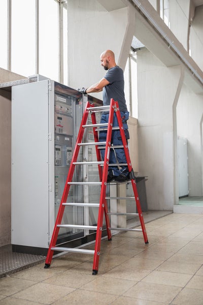 Handwerker auf einer Stufenleiter bei der Arbeit an einem Schaltschrank in einem Industriebetrieb