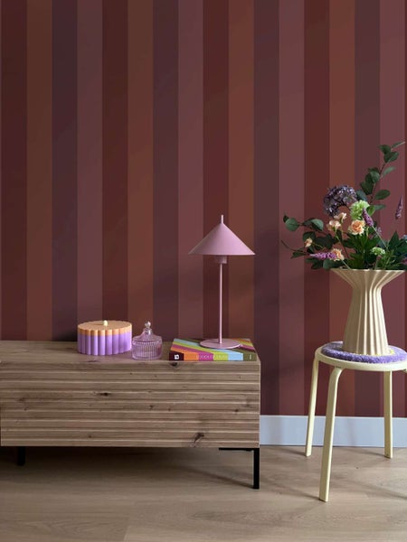Vliestapete mit vertikalem Streifenmuster in einer Wohnraumgestaltung mit Holzkommode, Lampe und einer Blumenvase auf einem Hocker.