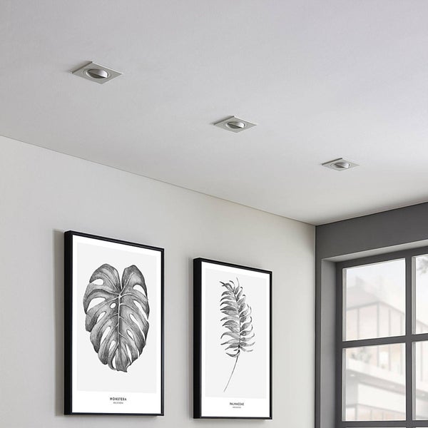 Drei quadratische Einbaustrahler aus Metall in einer weißen Decke über zwei gerahmten botanischen Bildern in einem hellen Raum.