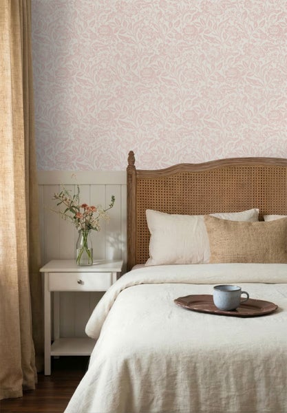 Schlafzimmer mit floraler Tapete in Rosa, Bett mit geflochtenem Kopfteil, weißem Nachttisch und beigen Vorhängen.