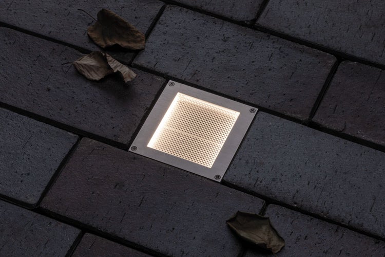 Quadratischer LED-Bodeneinbaustrahler aus Edelstahl in dunklem Pflasterstein, eingeschaltet mit warmem Licht.
