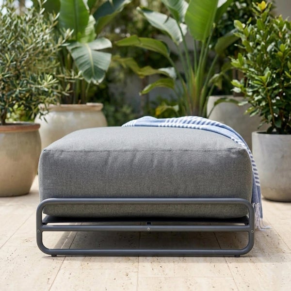 Moderner Gartenhocker mit Metallgestell und dickem Polster auf einer hellen Terrasse umgeben von mediterranen Kübelpflanzen.