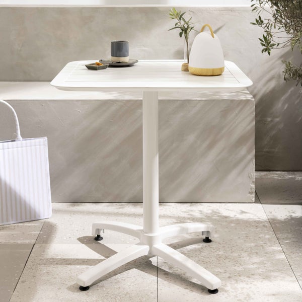 Weißer Bistrotisch mit quadratischer Leistenplatte und Mittelfuß auf einer hellen, minimalistischen Terrasse vor einer Steinwand.