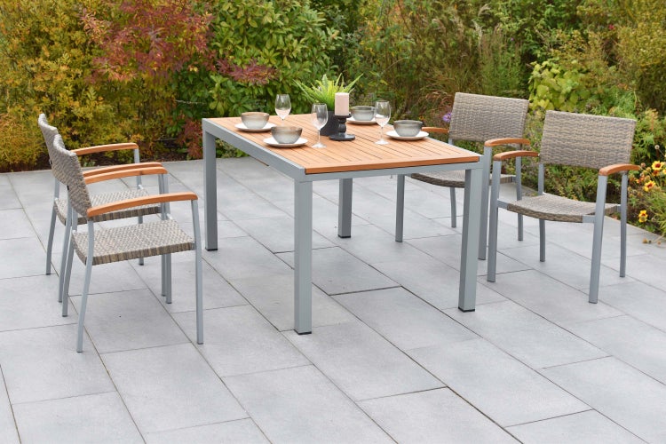 Modernes, helles Gartentischset mit einem Holztisch und vier Geflechtstühlen auf einer Terrasse aus Steinplatten umgeben von Pflanzen.