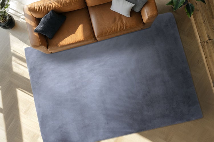 Ein unifarbener Wohnzimmerteppich mit weicher Oberfläche, platziert vor einem Ledersofa auf einem hellen Holzboden mit Fischgrätmuster.