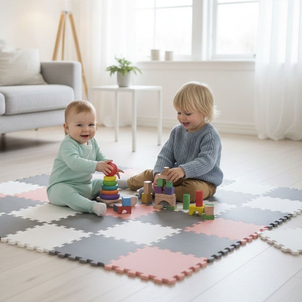 Zwei Kinder spielen auf einer bunten Puzzle Spielmatte aus Schaumstoff mit Holzspielzeug in einem hellen, modern eingerichteten Wohnzimmer.