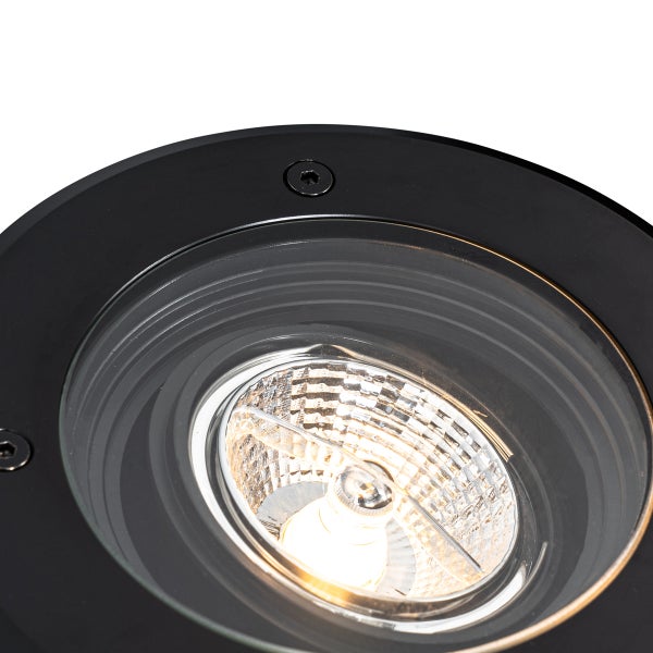 Nahaufnahme eines runden Bodeneinbaustrahlers mit schwarzem Rahmen, Glasabdeckung und eingeschaltetem Leuchtmittel.