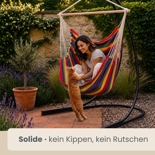 Frau sitzt in einem bunten Hängesessel mit Gestell im Garten und spielt mit einer Katze auf einer Terrasse aus Steinplatten.