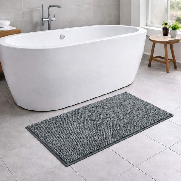 Graue Badematte mit Noppenstruktur vor einer weißen freistehenden Badewanne in einem modernen Badezimmer mit grauen Fliesen.