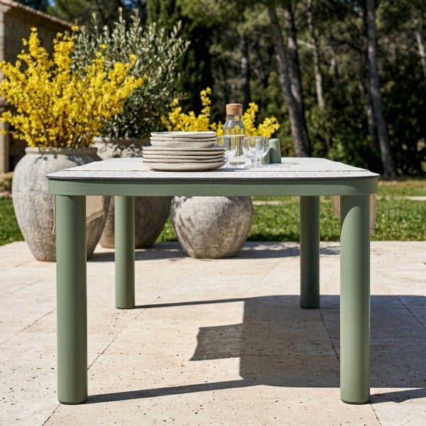 Moderner Gartentisch mit grünen Beinen und heller Steinplatte auf einer sonnigen Terrasse mit großen Pflanzgefäßen und Geschirr.