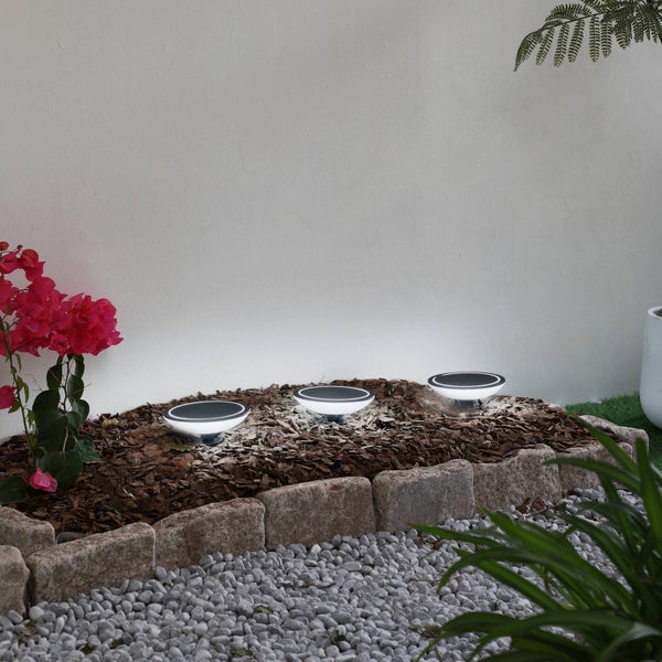 Drei runde Solar-Außenleuchten in einem Gartenbeet mit Rindenmulch, eingefasst von Natursteinen und umgeben von Pflanzen.
