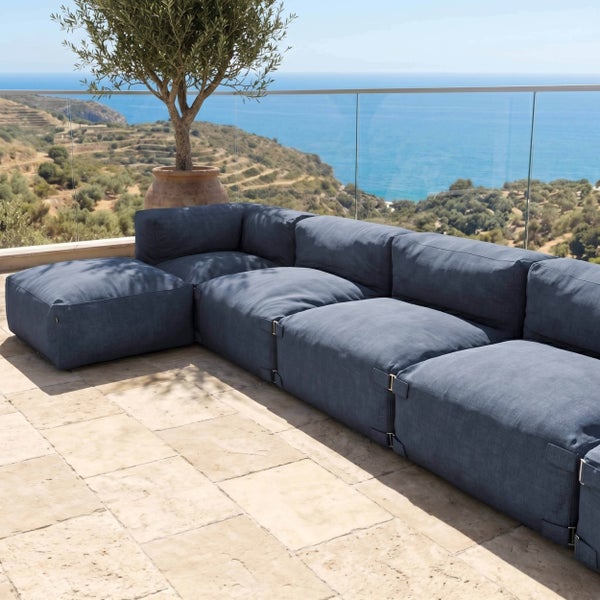 Modulares Outdoor-Sofa aus blauem Stoff auf einer hellen Steinterrasse mit Glasgeländer und Meerblick.