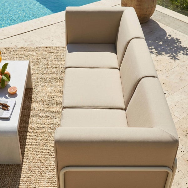 Modernes 3-Sitzer Gartensofa mit beigen Polstern auf einer Terrasse am Pool, neben einem Beistelltisch und einem gewebten Außenteppich.