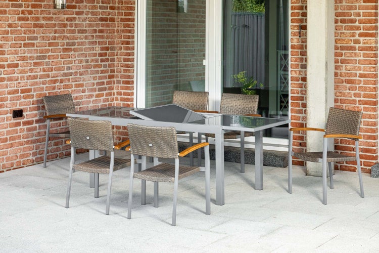 Helle, moderne Terrassen-Essgruppe mit grauem Metalltisch, Glasplatte und sechs passenden Stühlen mit Geflecht und Holzarmlehnen vor einer Ziegelwand.