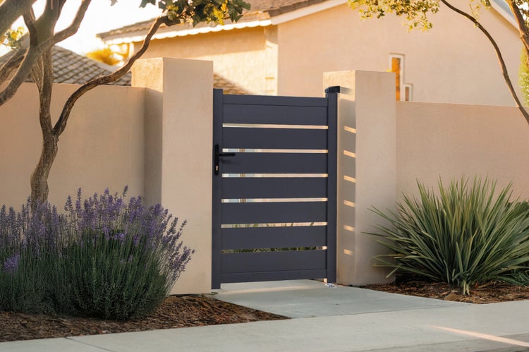 Modernes Gartentor aus Aluminium mit waagerechten Lamellen, montiert zwischen zwei hellen Mauerpfosten in einer Gartenanlage mit Pflanzen.