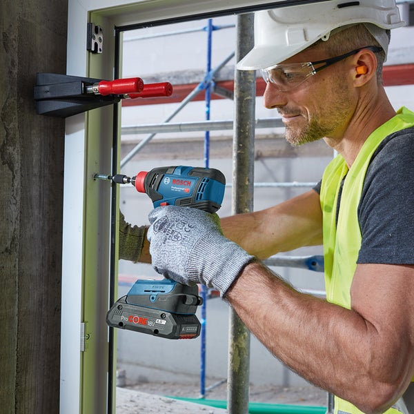Ein Mann mit Schutzbrille, Bauhelm und Sicherheitsweste schraubt mit einem Akku-Schlagschrauber eine Schraube in einen Fensterrahmen. Eine Fensterrahmenmontagehilfe ist befestigt. Bosch Logo.