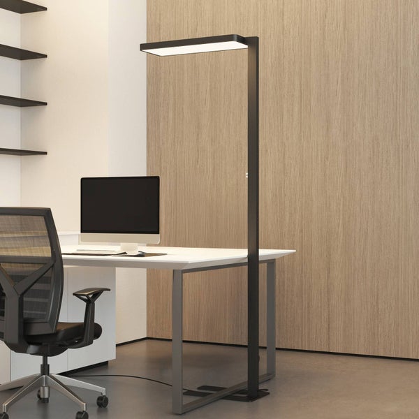 Moderne schwarze LED-Stehleuchte an einem Arbeitsplatz mit weißem Schreibtisch, Monitor und Bürostuhl in einem hellen Büro.
