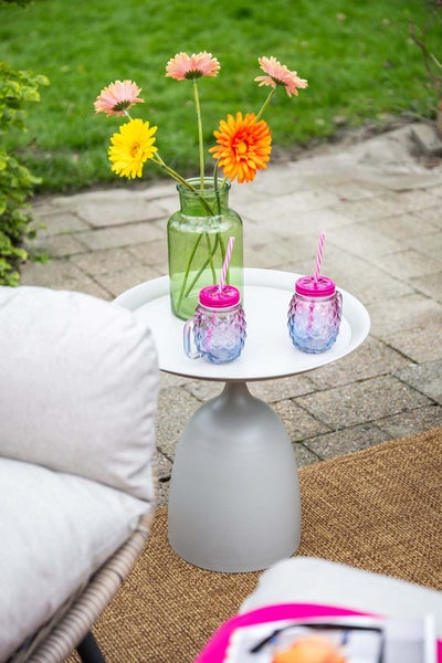 Runder Beistelltisch für den Garten mit einer Glasvase, Blumen und Trinkgläsern auf einer Terrasse mit Teppich.