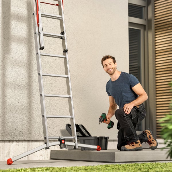 Mann kniet neben einer Leiter und einem Werkzeugkoffer und hält einen Akkuschrauber.