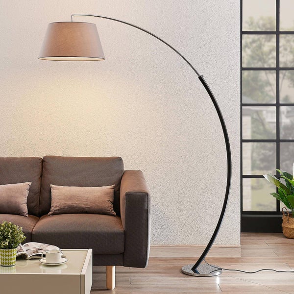 Moderne Bogenlampe mit Stoffschirm neben einem braunen Sofa in einem hellen Wohnzimmer mit Fenster.