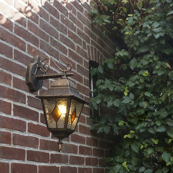 Klassische Außenwandleuchte im Laternendesign aus Metall mit strukturiertem Glas, montiert an einer Ziegelwand neben Laub.