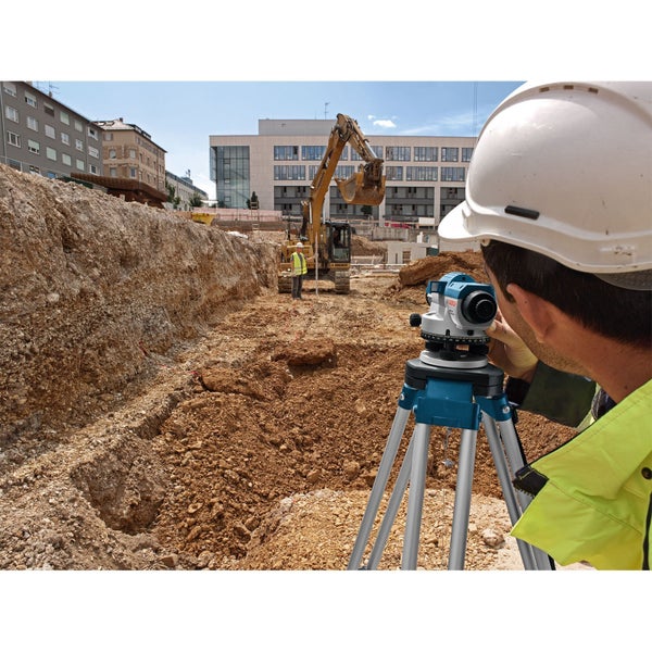Ein Bauarbeiter benutzt ein Nivelliergerät auf einer Baustelle, im Hintergrund ein Bagger.