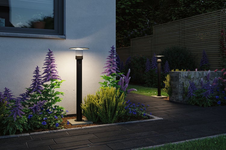 Zwei moderne schwarze Wegeleuchten beleuchten einen gepflasterten Gartenweg neben einem Blumenbeet und einer Hauswand in der Dämmerung.