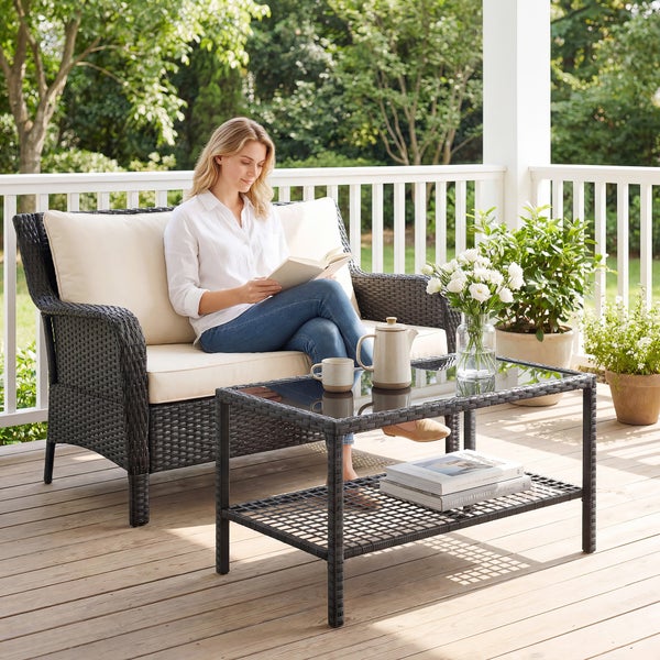 Gartenmöbel-Set aus dunklem Polyrattan mit 2-Sitzer Sofa, hellen Kissen und Couchtisch mit Glasplatte auf einer Holzterrasse.