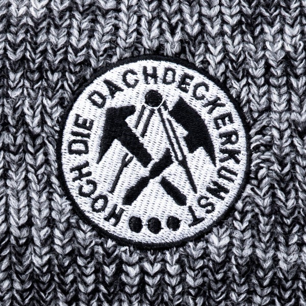 Besticktes Emblem mit Zunftlogo der Dachdecker und Schriftzug Hoch die Dachdeckerkunst auf grob gestricktem Stoff.