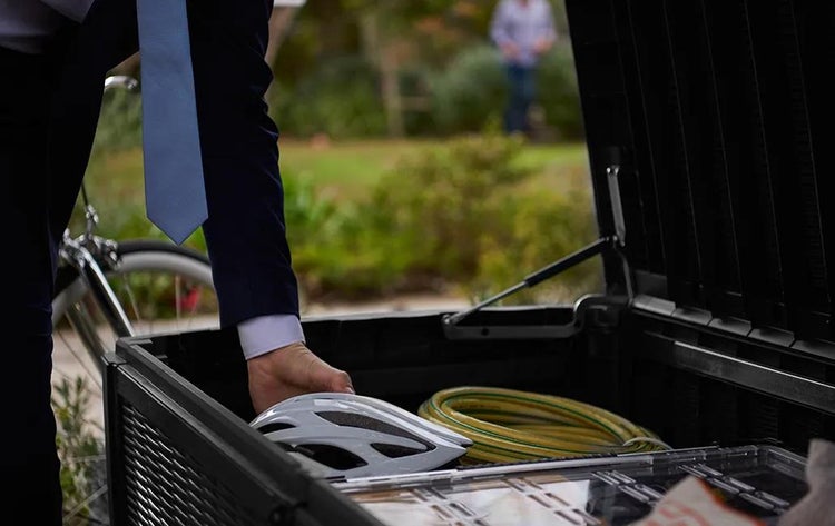 Eine Person legt einen Fahrradhelm in eine geöffnete Gartenbox aus Kunststoff, in der ein Gartenschlauch und eine Sortimentsbox verstaut sind.